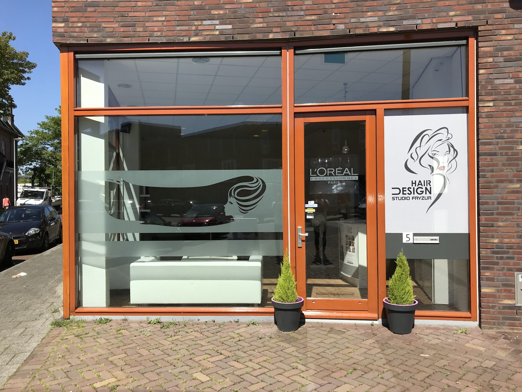 De etalage aan de Verbeetenstraat met oranje kozijnen, het logo op het raam en twee plantjes bij de deur