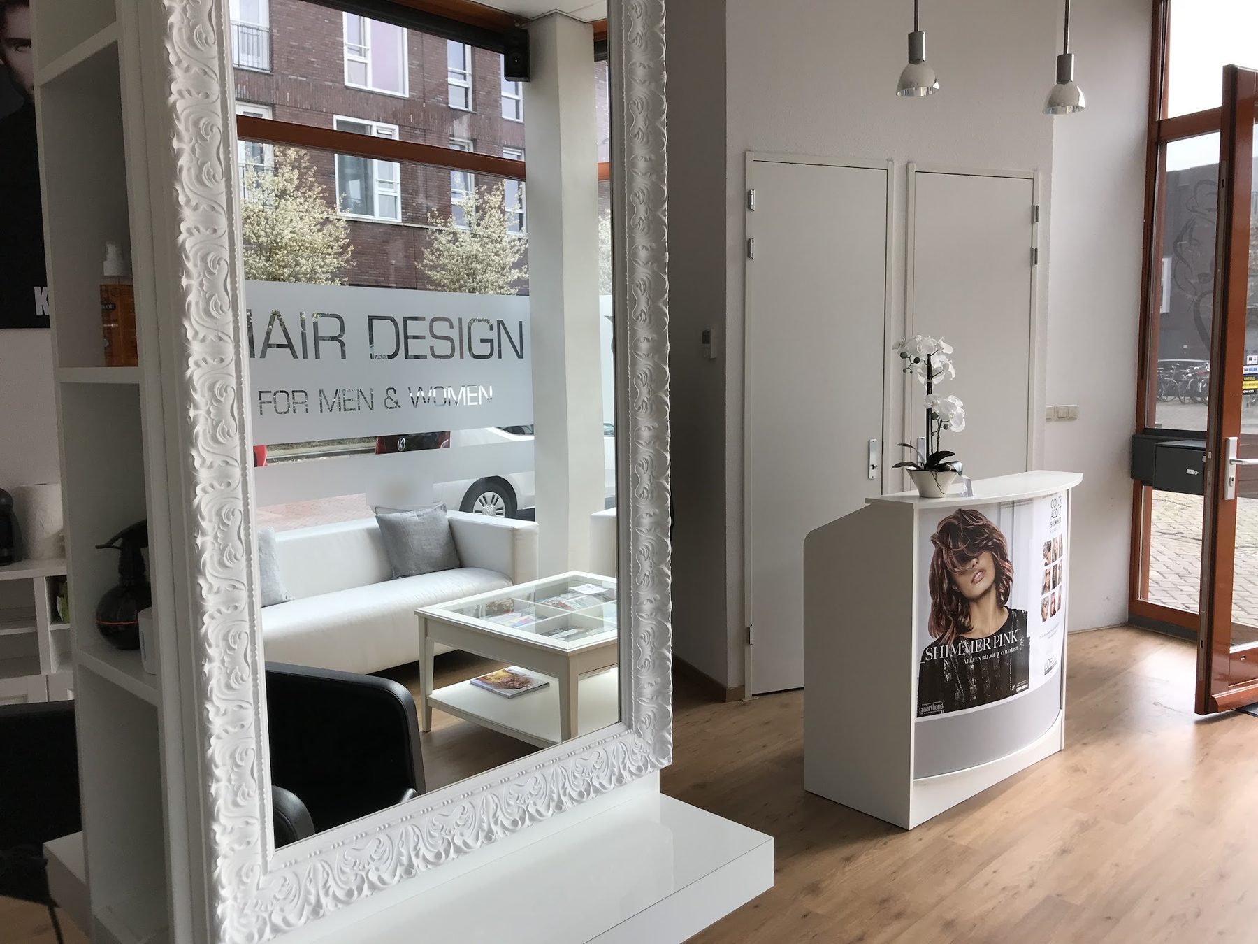 Een grote witte spiegel met bewerkte lijst, daarachter de balie en HAIR DESIGN FOR MEN & WOMEN op het raam