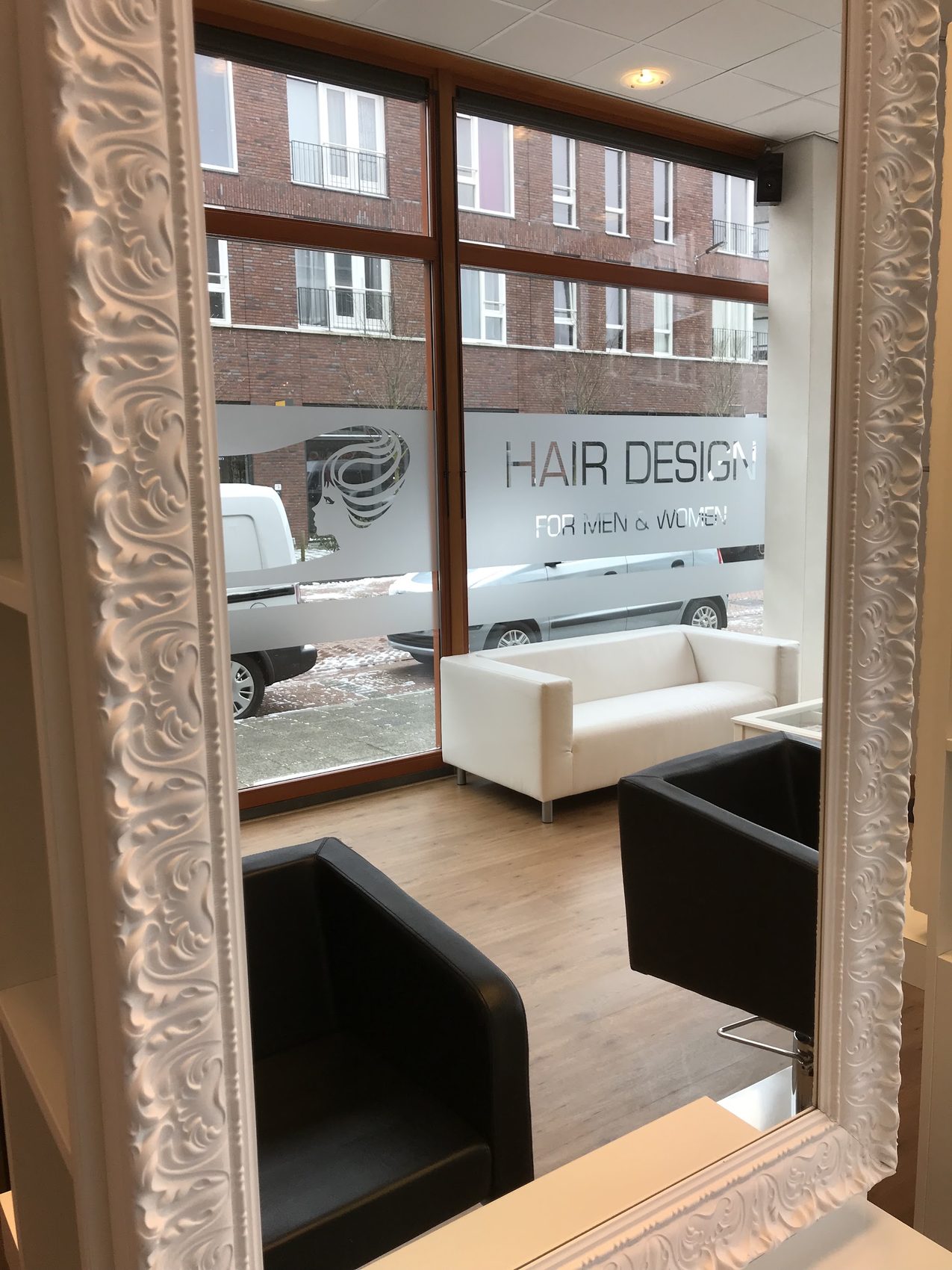De salon gezien langs een witte, bewerkte spiegel: zwarte stoelen, een witte bank en HAIR DESIGN op het raam